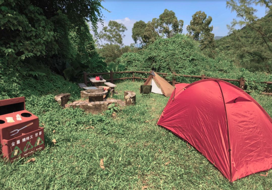 一个维护良好的AFCD营地,多个帐篷搭建在平整地面,前方有混凝土烧烤炉,后方有基本厕所设施,入口处有木质指示牌,四周为郁郁葱葱的郊野公园植被,天空阴沉,展现有组织且易于到达的露营环境